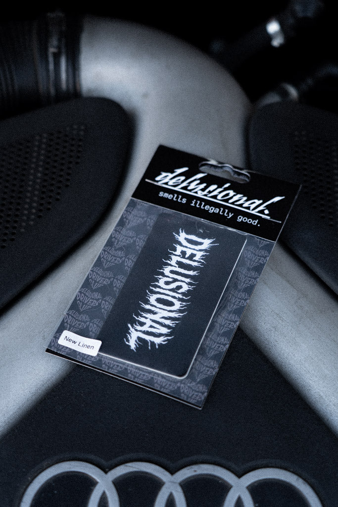 Duftbaum Produktbild von Delusional, mit Verpackung auf einer Motorhaube